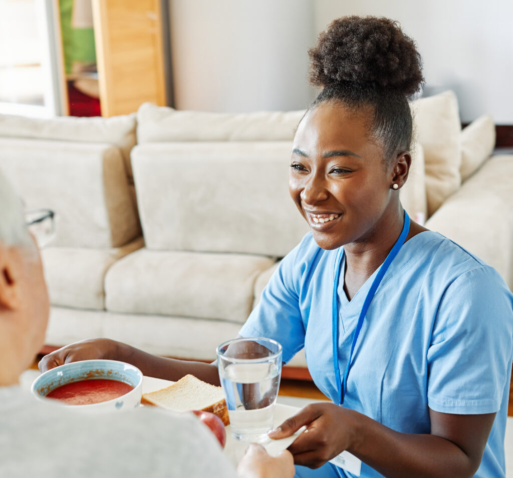 Professional home healthcare caregiver serving a meal and water to a senior adult in a comfortable home setting.